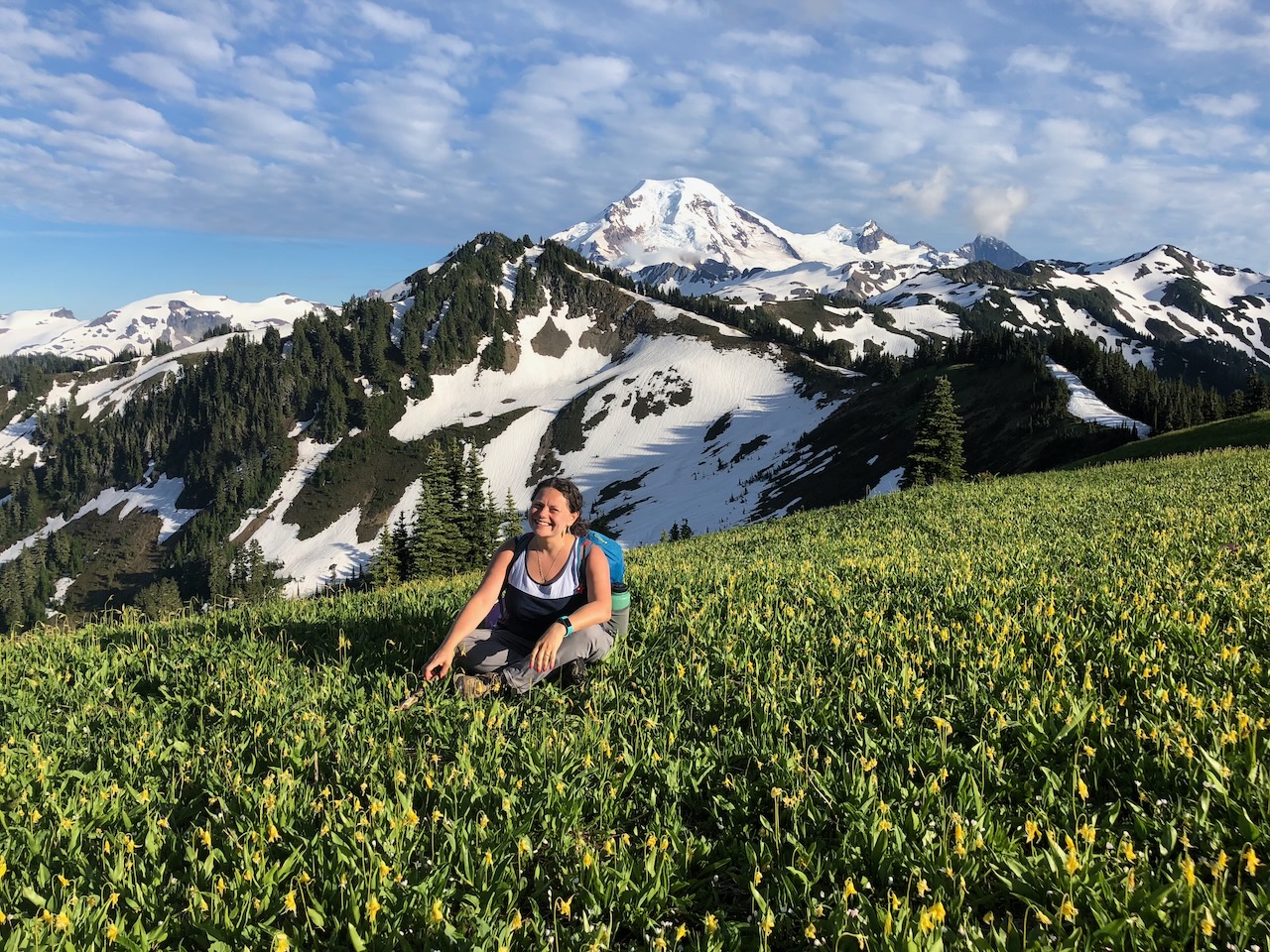 Meryl smiling in a wildflower meadow with snow-capped peaks behind her